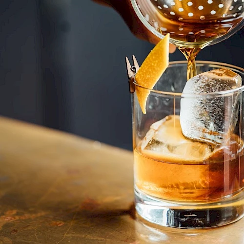 A whiskey glass with ice, a citrus twist garnish, and a whiskey being poured over it, on a wooden bar.