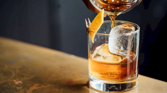 A whiskey glass with ice, a citrus twist garnish, and a whiskey being poured over it, on a wooden bar.
