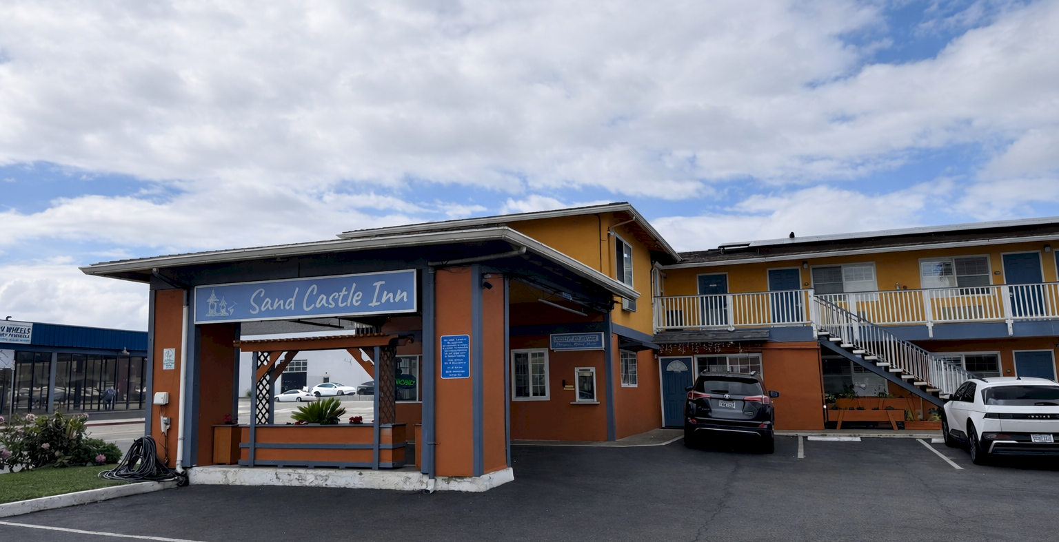 A small motel with orange and tan exterior, a sign reading &ldquo;Sand Castle Inn,&rdquo; a drive-up entrance, parked cars, and a fenced balcony on a sunny day.