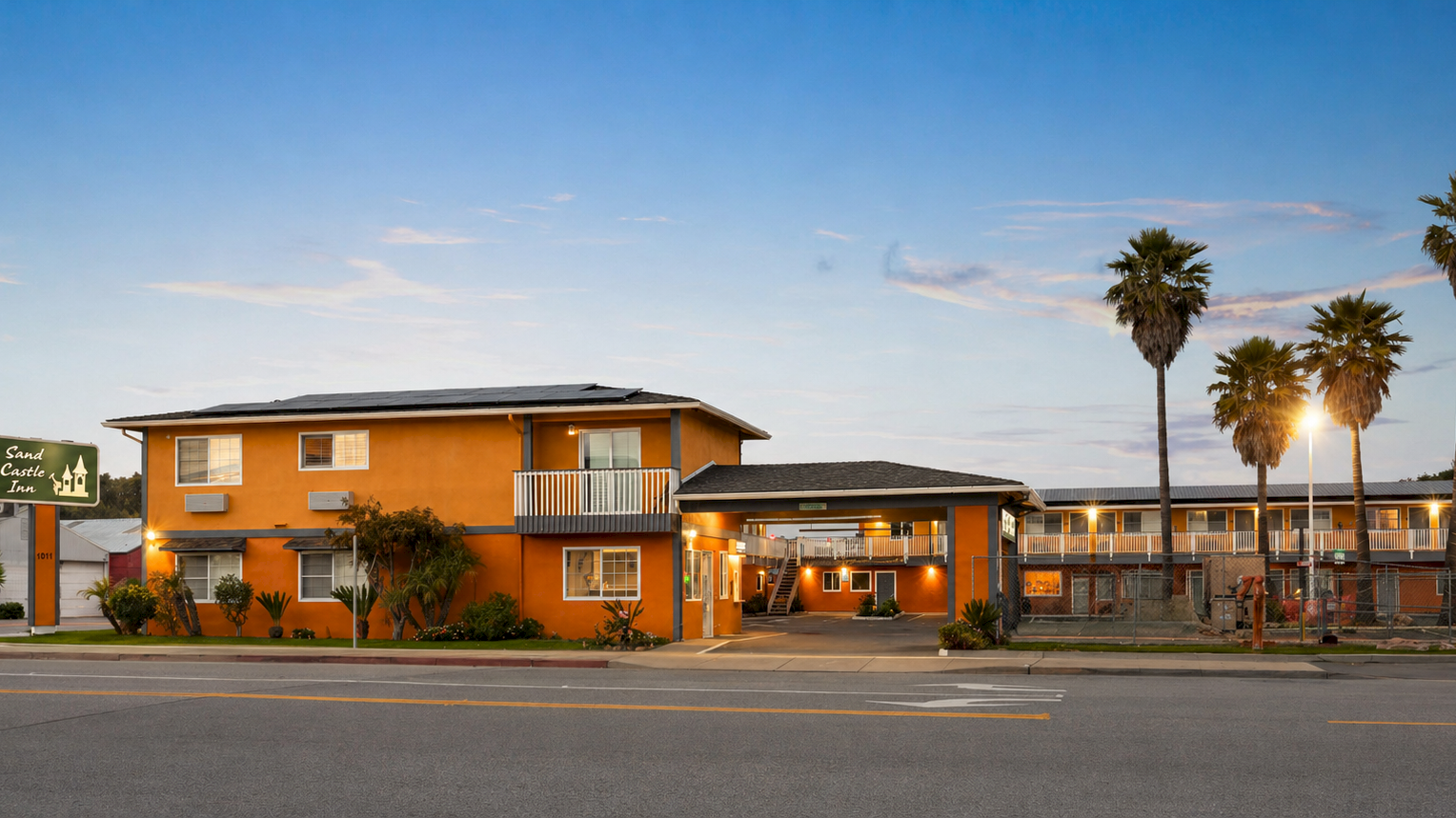 A yellow-orange motel building with balconies and palm trees at dusk, with &ldquo;Sand Castle Inn&rdquo; signage by the road.