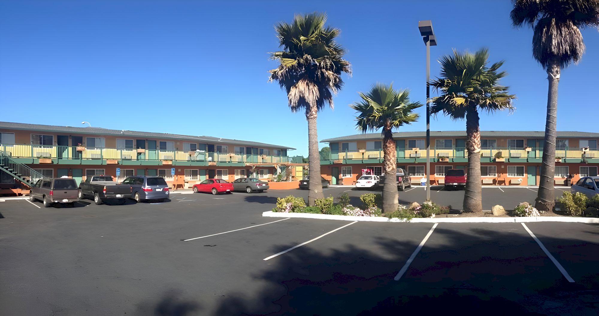 A motel parking lot with a two-story beige building, several palm trees, parked cars, and clear blue sky.