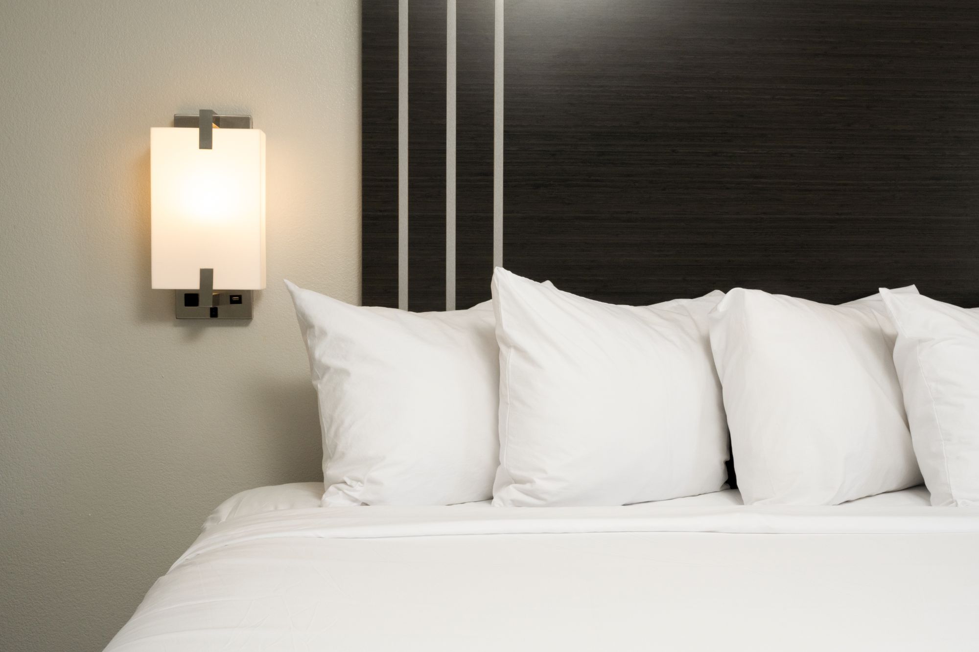 A neatly made bed with white pillows and sheets, a modern wall lamp on the left, dark headboard with vertical white lines, calm hotel vibe.