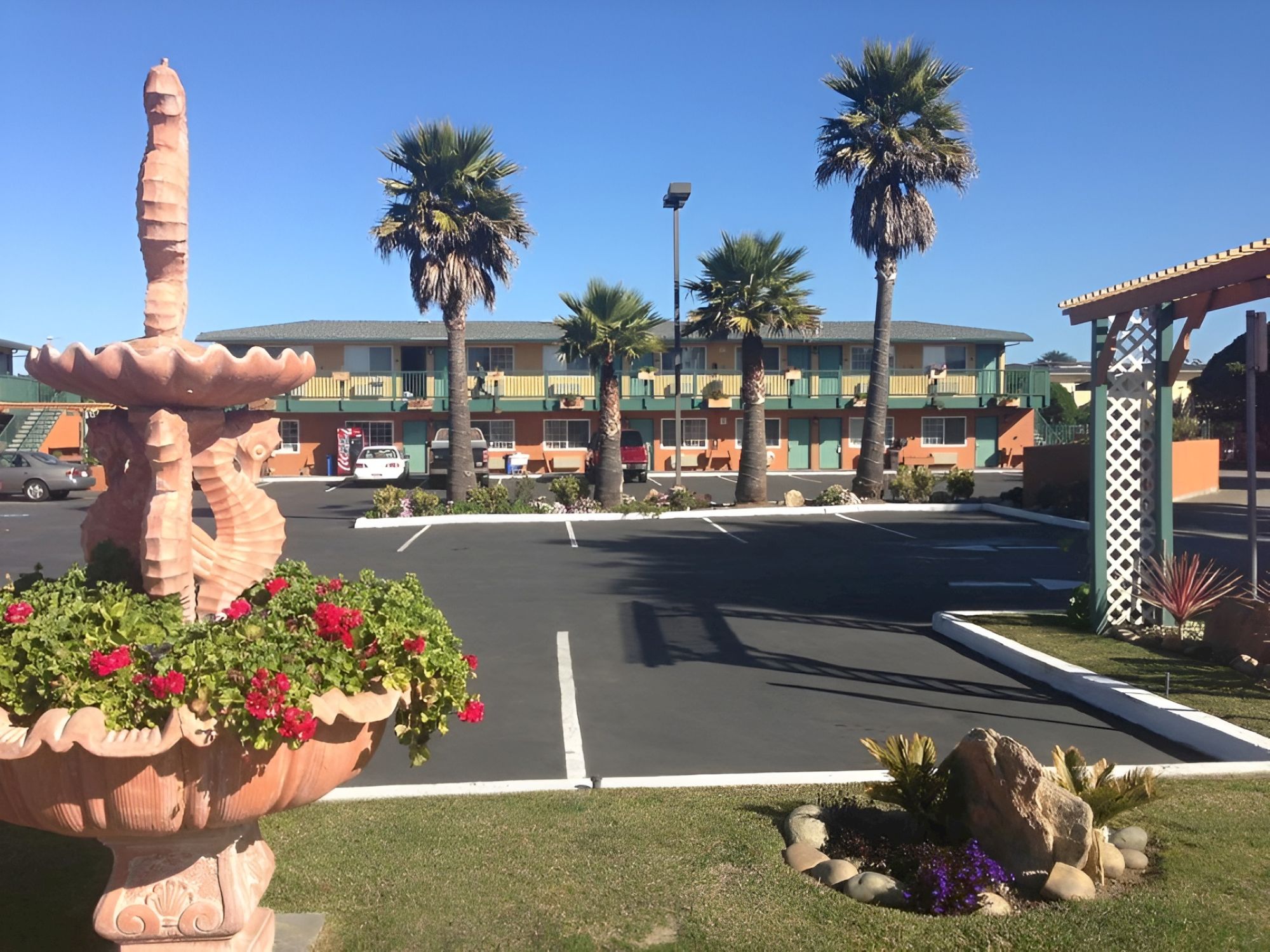 A motel-style parking lot with palm trees, a stone fountain in the foreground, and a two-story building in the background, under a clear blue sky.