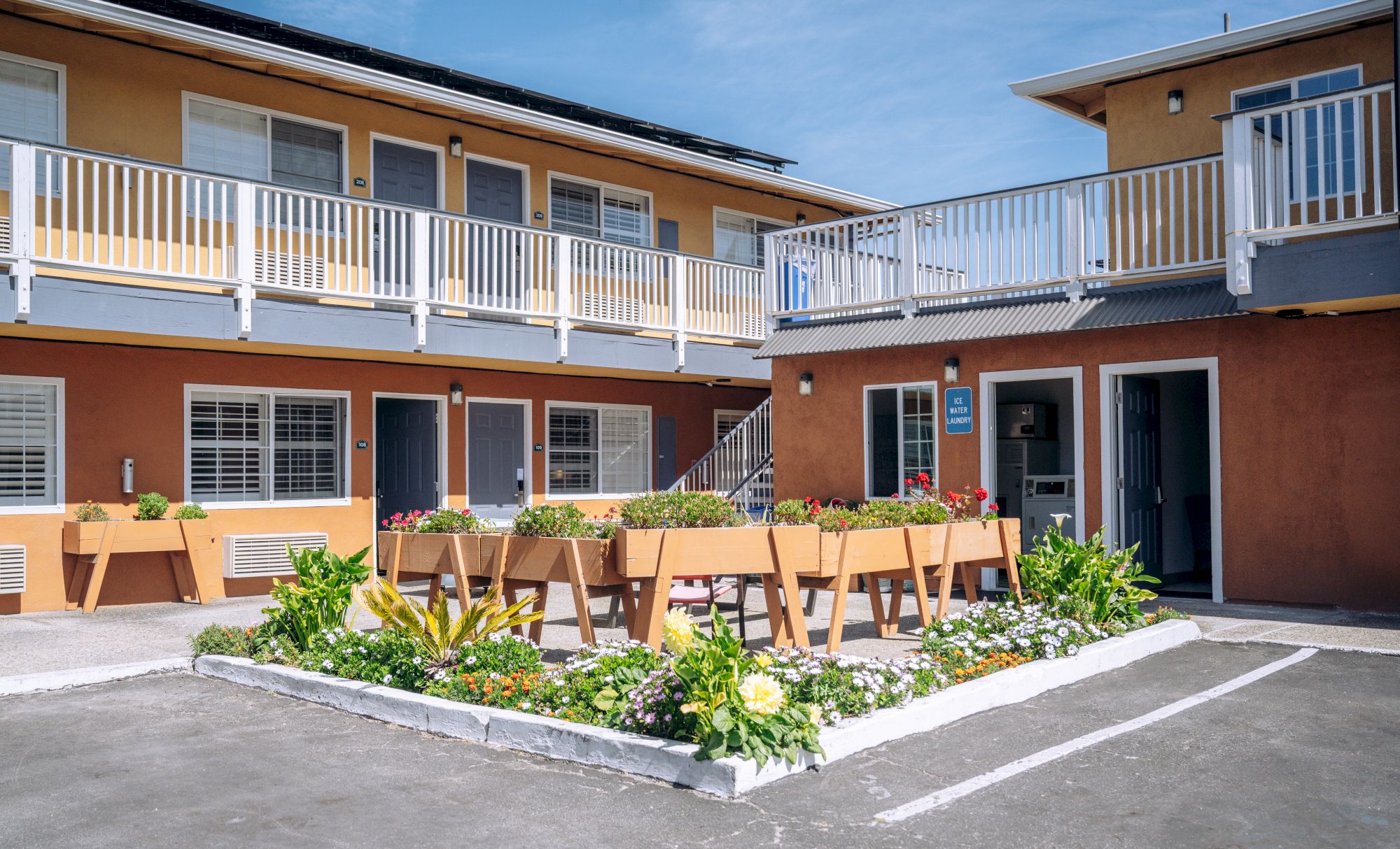 Two-story motel courtyard with orange walls, white railings, outdoor planters, and a long table set for guests in the center, under a sunny sky.