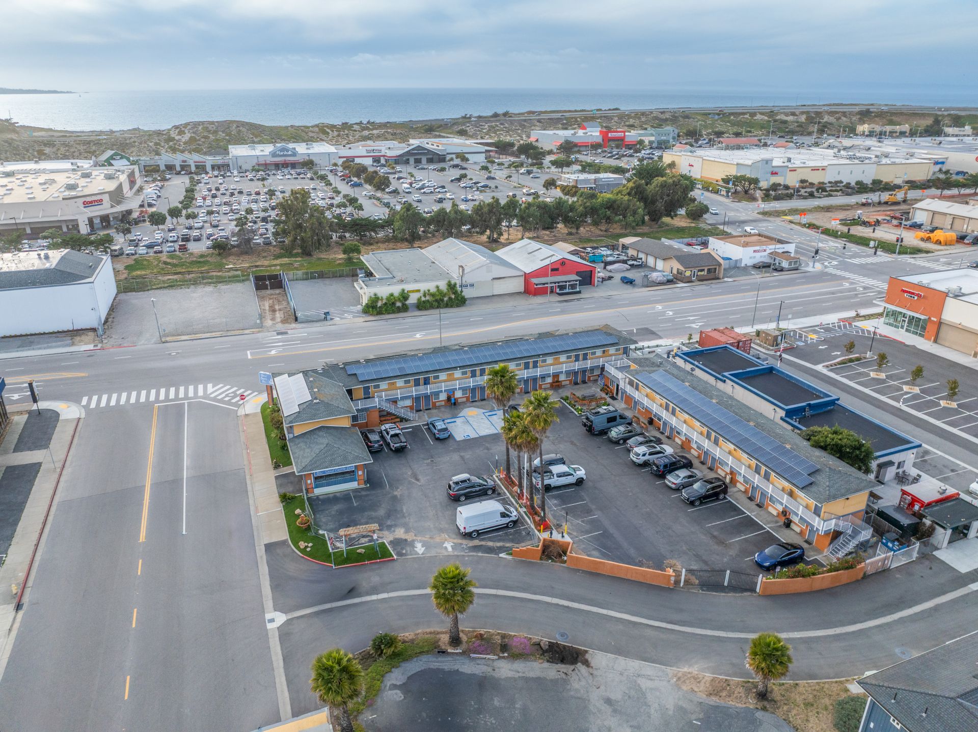 Aerial view of a roadside strip with a small motel or diner, parking lot, and a few palm trees; surrounding commercial and residential blocks stretch to the horizon.