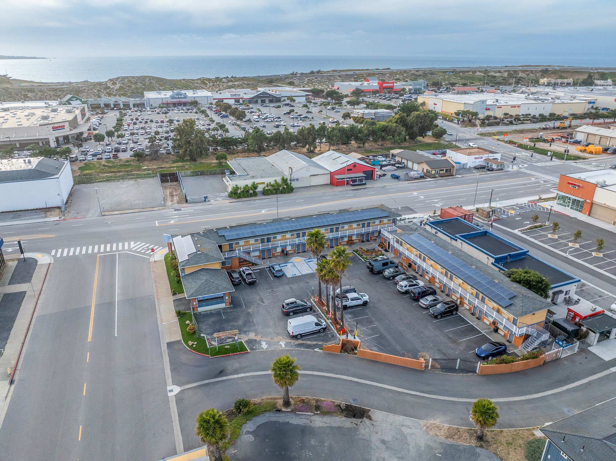Aerial view of a roadside strip with a small motel or diner, parking lot, and a few palm trees; surrounding commercial and residential blocks stretch to the horizon.