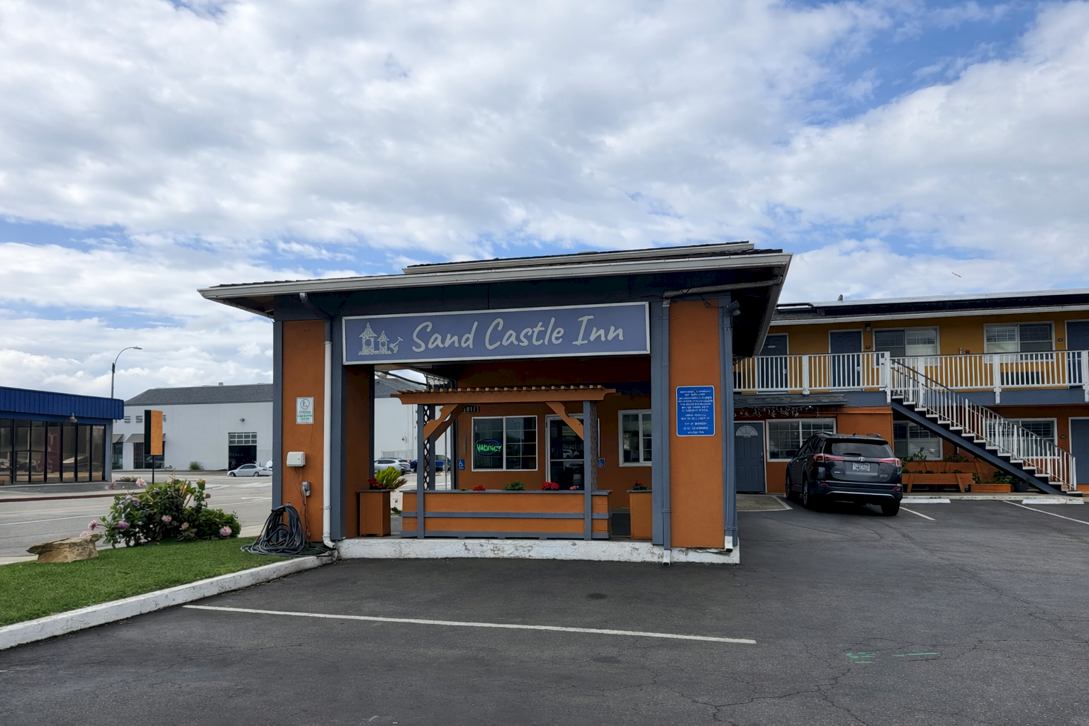 A roadside inn with a wooden-like entrance sign that reads &ldquo;Sand Castle Inn,&rdquo; covered parking, and a blue sky with clouds, underlit by sunlight.