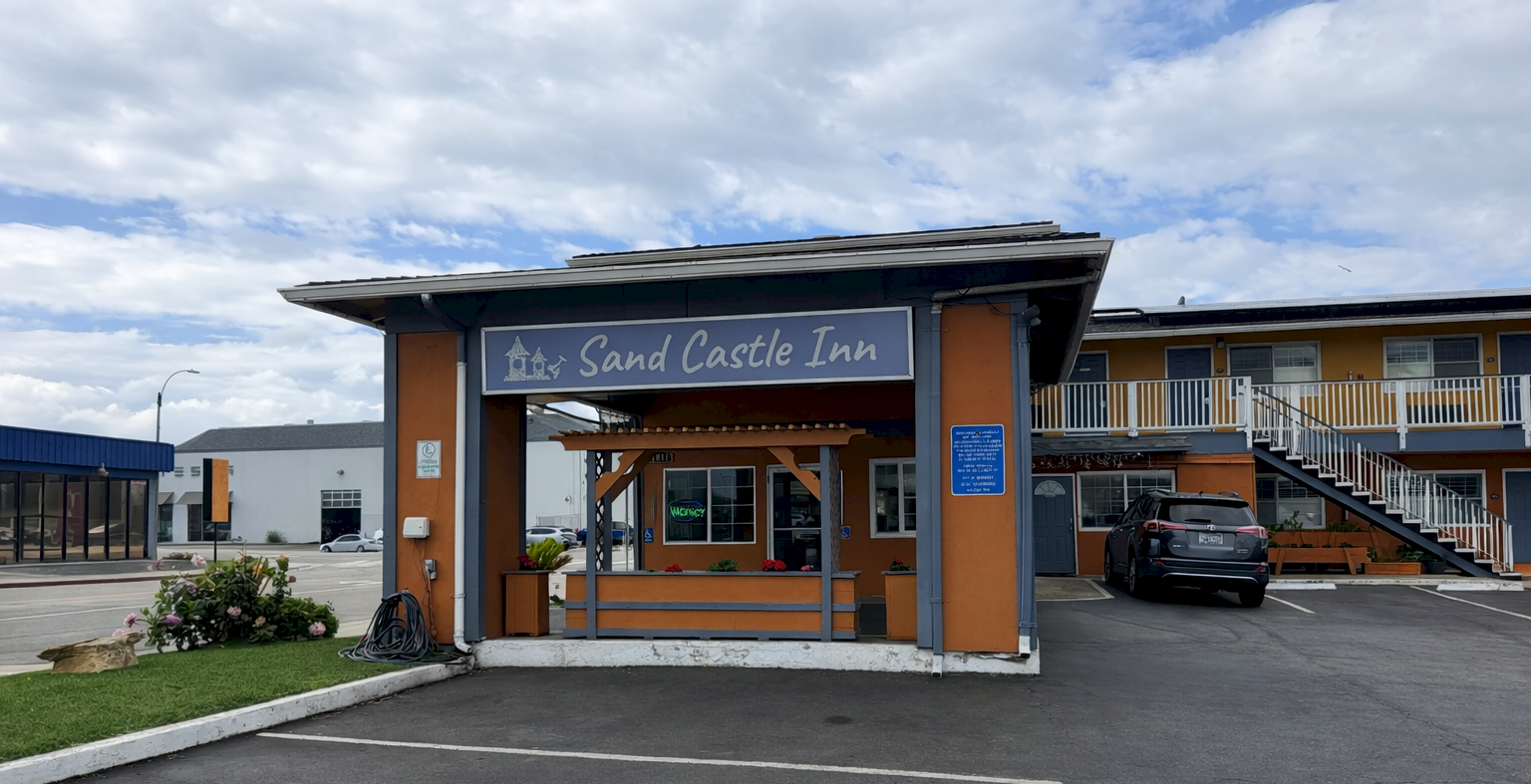 A roadside inn with a wooden-like entrance sign that reads &ldquo;Sand Castle Inn,&rdquo; covered parking, and a blue sky with clouds, underlit by sunlight.