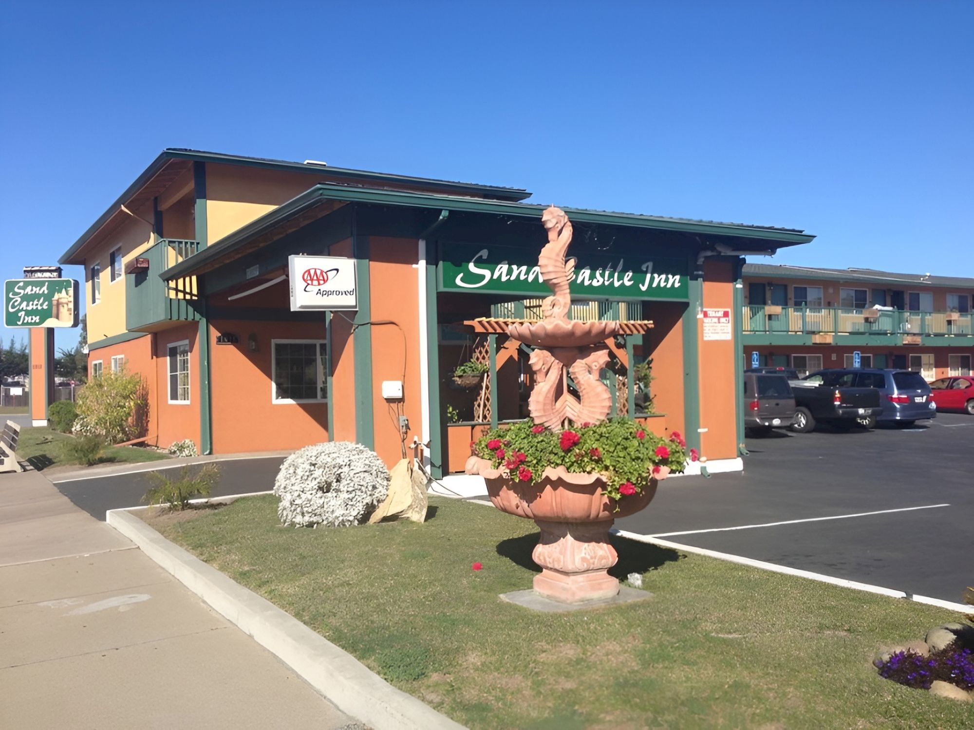 A sunny, small-town storefront with a &ldquo;Sweet music shop&rdquo; sign and a large statue planter in front of the entrance, nearby parking and modern building.