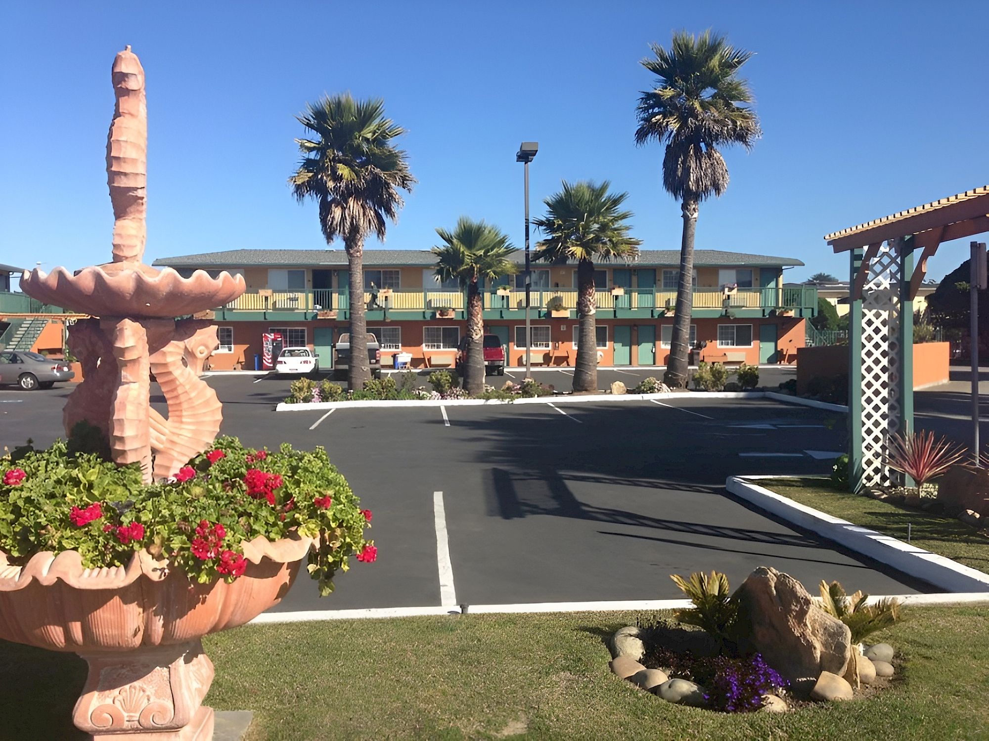 A motel parking lot with palm trees, flower planters, and a pink sculpted fountain in the foreground, under a clear blue sky.