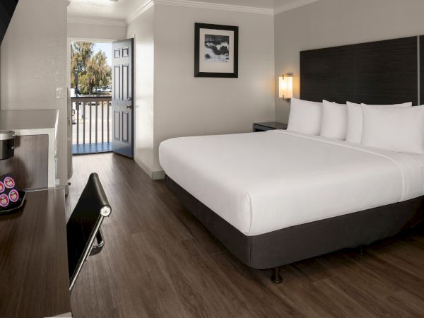 A modern hotel room with a large white bed, dark headboard, nightstands, a balcony door, and a small bench or luggage rack at the foot of the bed. The space is tidy and bright.