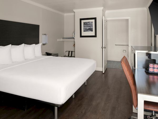 A modern hotel room with a large white bed, dark headboard, desk, chair, and a doorway leading to another area.