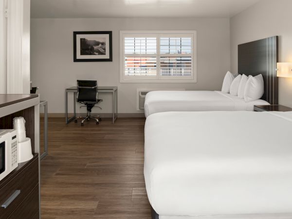 A hotel room with three white beds, a desk and chair, a window with blinds, framed wall art, and a small kitchenette corner nearby.