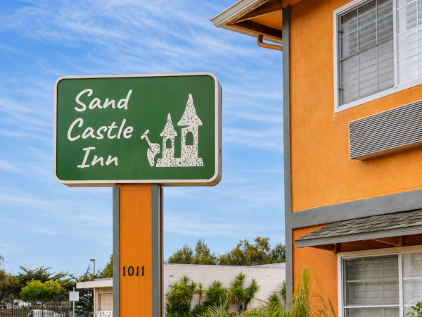 A green sign reads &ldquo;Sand Castle Inn&rdquo; with a small castle graphic, beside an orange building under a blue sky.