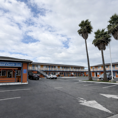 A beachfront motel with a single-story orange-and-blue building, palm trees, and a mostly empty parking lot under a partly cloudy sky.