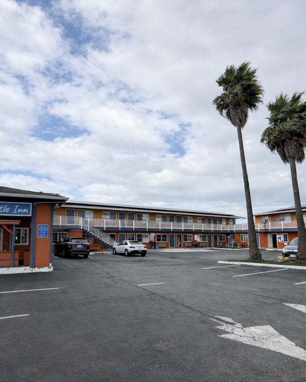 A beachfront motel with a single-story orange-and-blue building, palm trees, and a mostly empty parking lot under a partly cloudy sky.