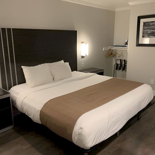 A tidy hotel room with a large bed, white linens, brown bed runner, dark headboard, nightstands, wall lights, and a small open closet area.