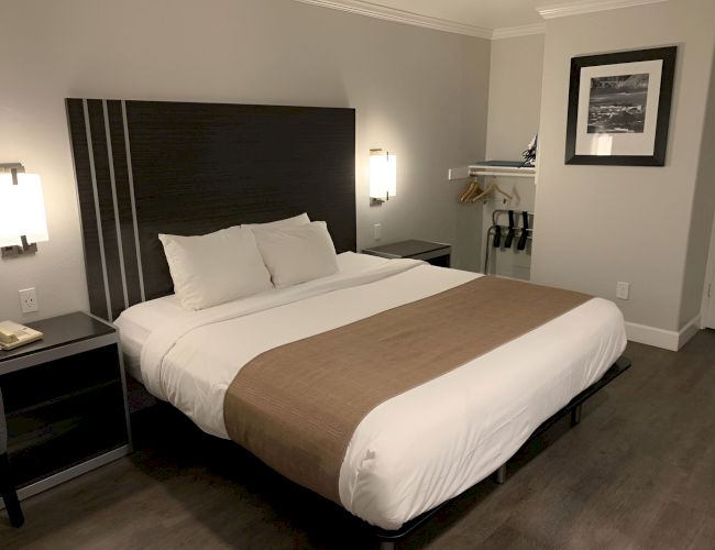 A neatly made hotel bed with white linens and a brown runner, dark headboard, nightstands, wall lamps, and a small open closet space.