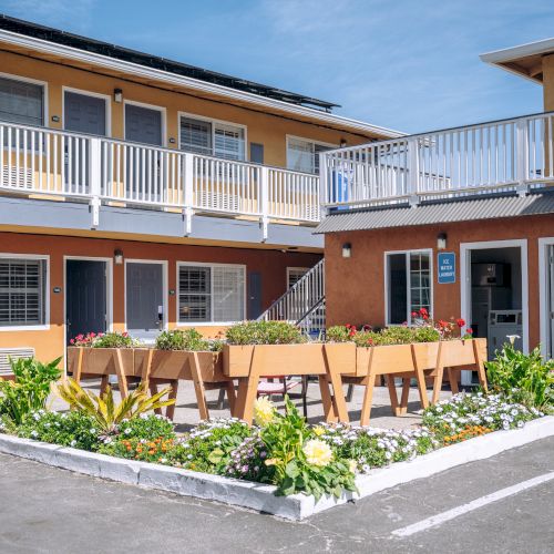 Outdoor motel-style courtyard with a two-story building, patio tables, and a small garden bed in the center, in a sunny, weekday scene.