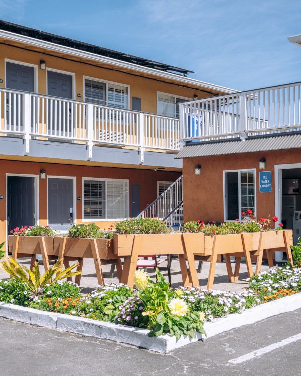 Outdoor motel-style courtyard with a two-story building, patio tables, and a small garden bed in the center, in a sunny, weekday scene.