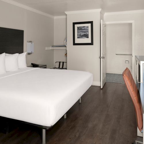 A modern hotel room with a large white bed, dark headboard, wooden flooring, small desk, chair, and a doorway to the bathroom.