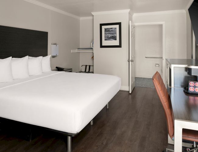 A modern hotel room with a large white bed, dark headboard, wooden flooring, small desk, chair, and a doorway to the bathroom.