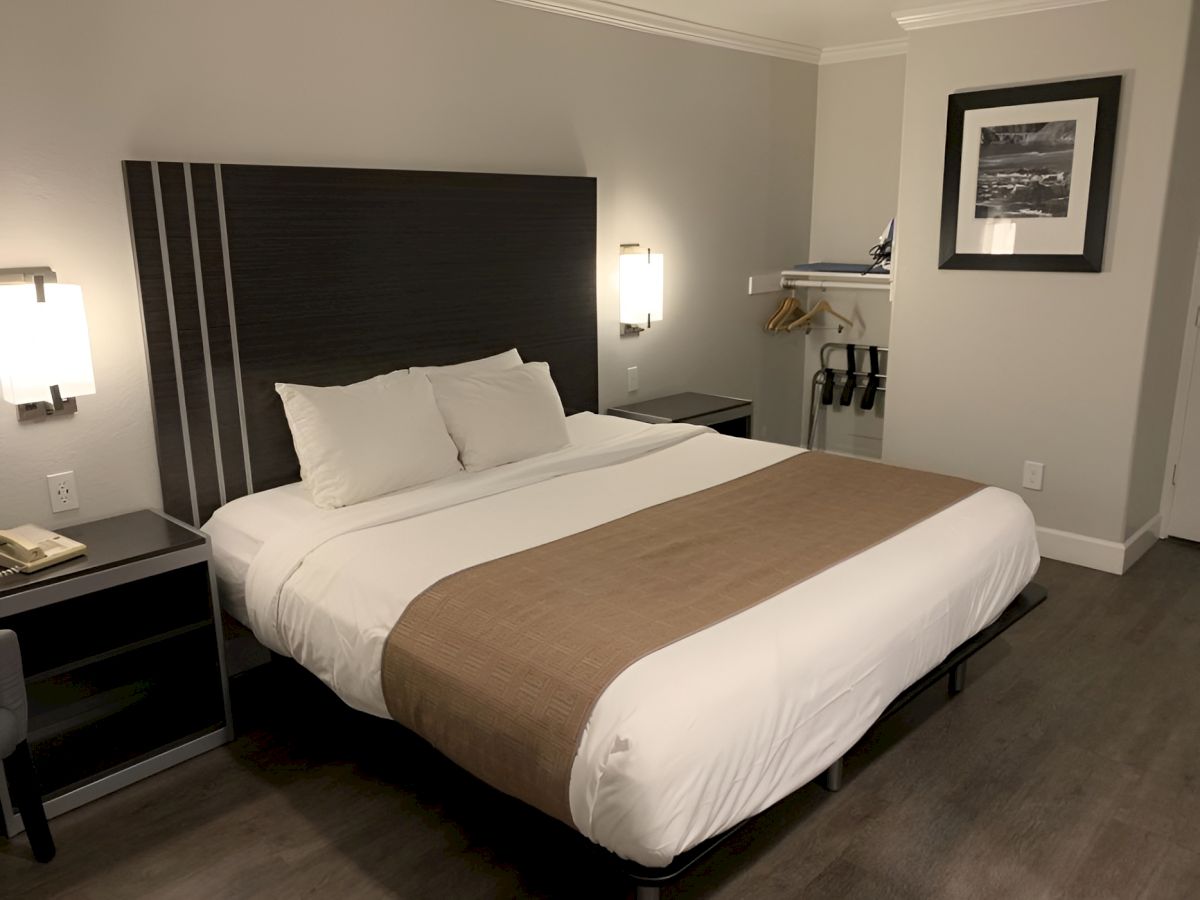 A modern hotel room with a large bed, white linens, brown throw, dark wood furniture, and a small desk area in a light gray/neutral palette.