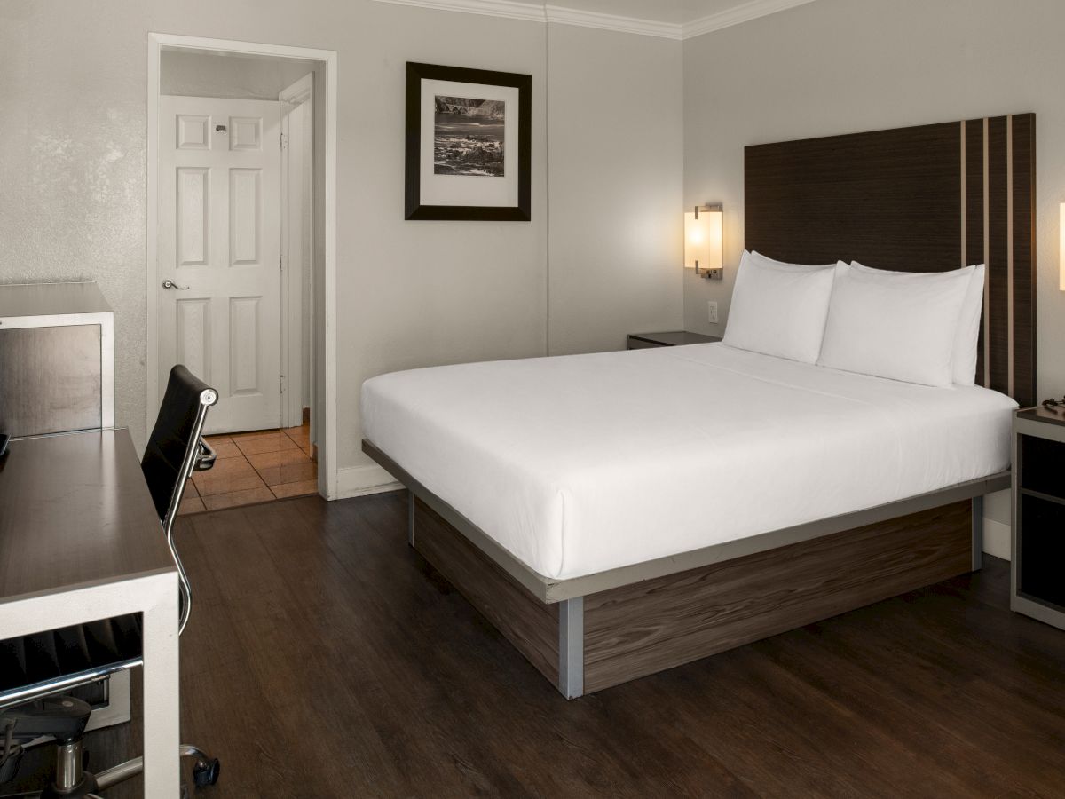 A tidy hotel room with a king bed, wooden headboard, two nightstands, desk, chair, and a framed picture on the wall, door open to a hallway.