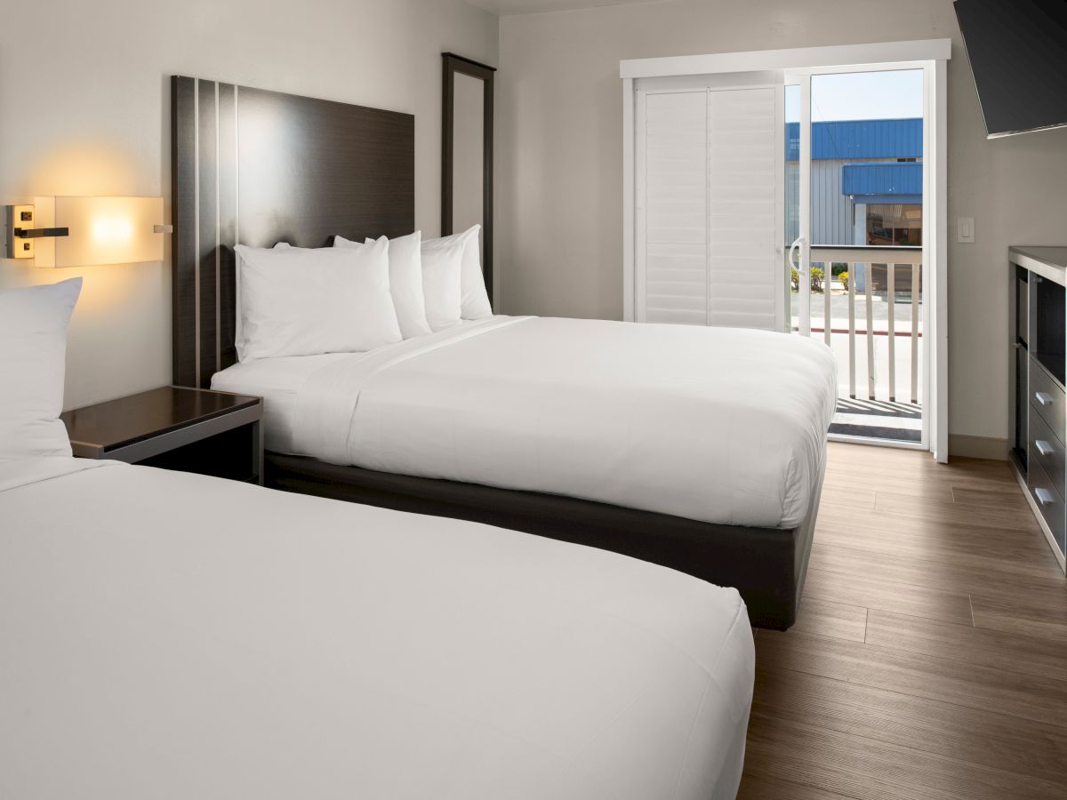 Two neatly made hotel beds with white linens, a nightstand, warm wall light, and a door to a balcony overlooking a sunny view.