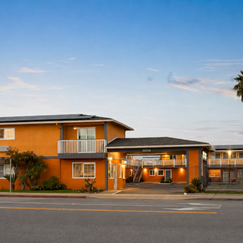 A retro motel with orange exterior, palm trees, and a lit welcome sign under a dusky sky, giving off a classic roadside vibe.