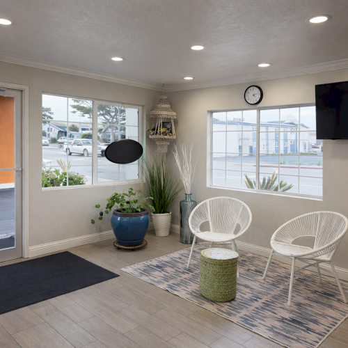 A modern living room with a light gray sofa, glass coffee table, TV on a dark wall, white round chair, plants, and large windows with a balcony.