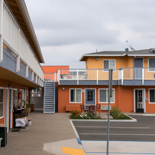 A small orange-and-white motel with a parking lot, two stories, rows of rooms, a sign, and a few trees in the background, under a cloudy sky.