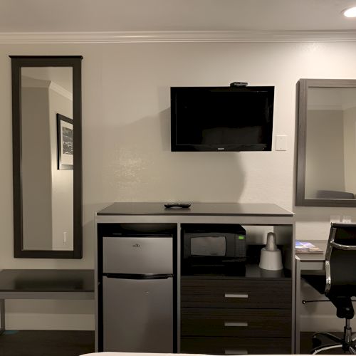 A modern hotel or office suite with a desk, chair, wall-mounted TV, artwork, and a large mirror, all in neutral tones, neat and contemporary.