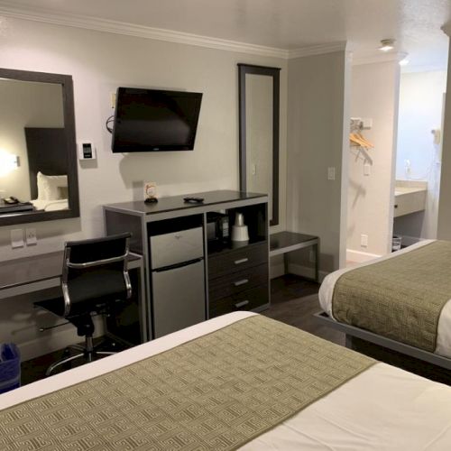 A hotel room with two beds, a desk, a wall-mounted TV, a mirror, a small fridge, and a chair; neutral decor with beige and gray tones.
