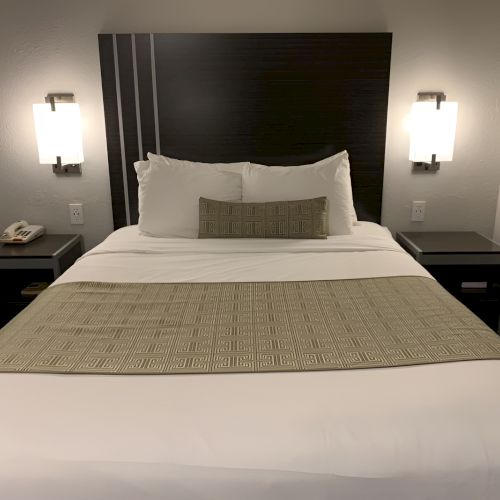 A neatly made bed with white linens and a beige runner, two nightstands with lamps flanking it, in a minimalist hotel room.