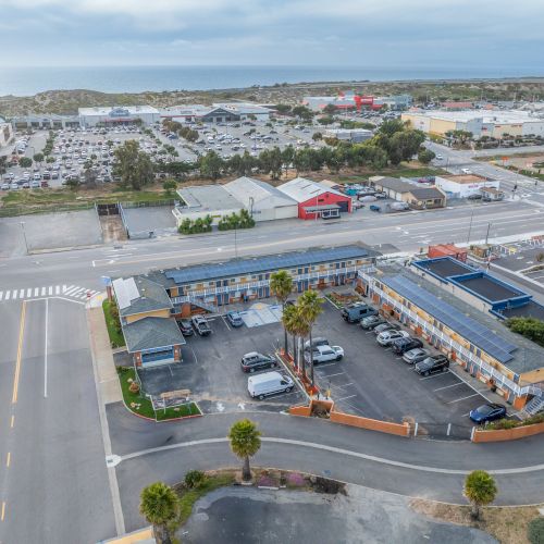 Aerial view of a parking lot and strip with a fast-food style restaurant, adjacent buildings, palm trees, and a distant commercial area.