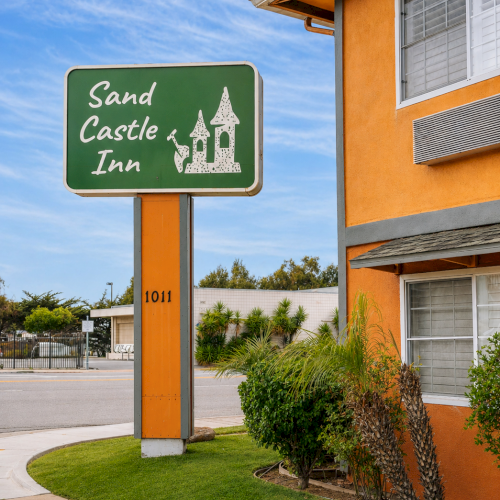 A sign reads &ldquo;Sand Castle Inn&rdquo; with a small castle graphic beside a two-story orange building, a sidewalk, and blue sky in the background.