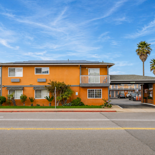 A mid-century orange motel with a carport hallway, palm trees, and a roadside sign along a sunny street.