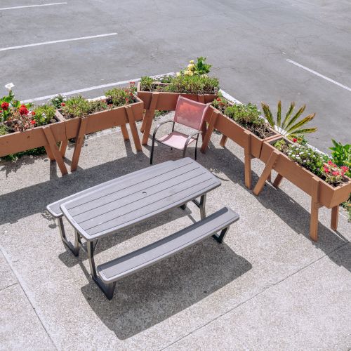 A small outdoor seating area with a curved wooden planter filled with flowers surrounds a gray picnic table and bench, placed on a concrete surface.