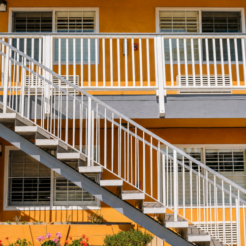A bright orange apartment building with white railings and stairs, windows with blinds, and potted plants along the ground adds charm.
