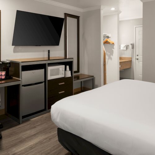 A modern hotel room with a desk, flat-screen TV, mini-fridge, and a bed; glimpses of a wardrobe and an open bathroom in the background.