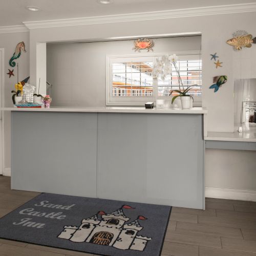 A small reception desk area with a light gray counter, door on the left, white walls, and ocean-themed wall decorations; a mural rug on the floor reads &ldquo;Send good vibes.&rdquo;