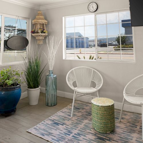 A cozy sunlit balcony with white wicker chairs, a woven stool, green plants in blue and white pots, and a rug by the windows overlooking a view.