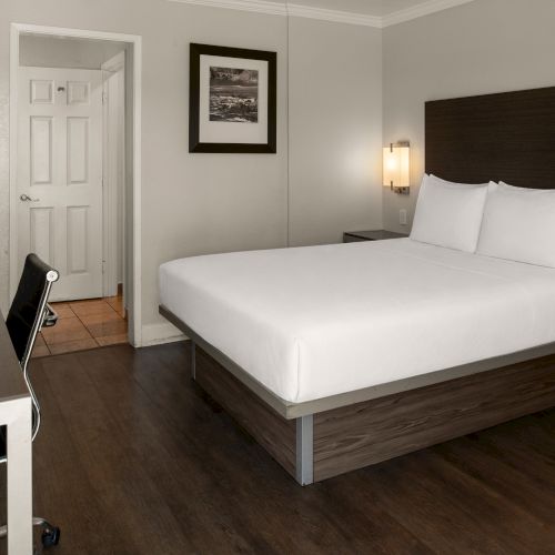 A tidy hotel room with a king bed, two nightstands, wall lamps, a desk and chair, framed art, and a door to a small hallway.