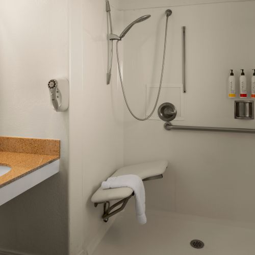 A modern hotel bathroom with a shower stall, sink with granite countertop, mirror, and a wall-mounted grab bar and amenities cabinet, all neat and clean.