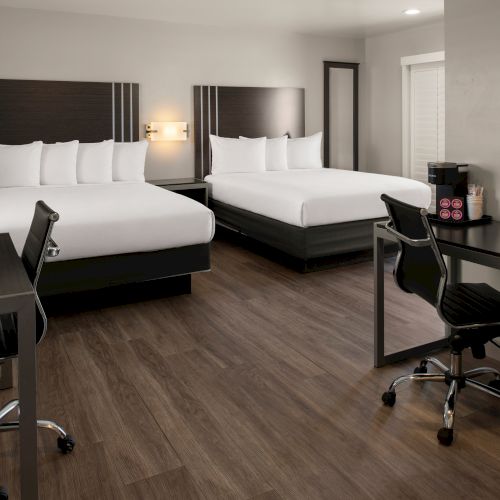 A neat hotel room with two large beds, a desk and chair, a dresser, and a mirrored wall, all in a modern, neutral-toned setting.