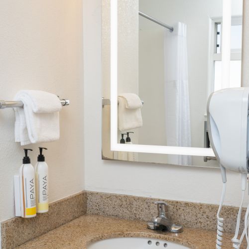 A bathroom vanity with a granite countertop, under-mount sink, illuminated framed mirror, mounted hairdryer, and stocked toiletries on the counter.