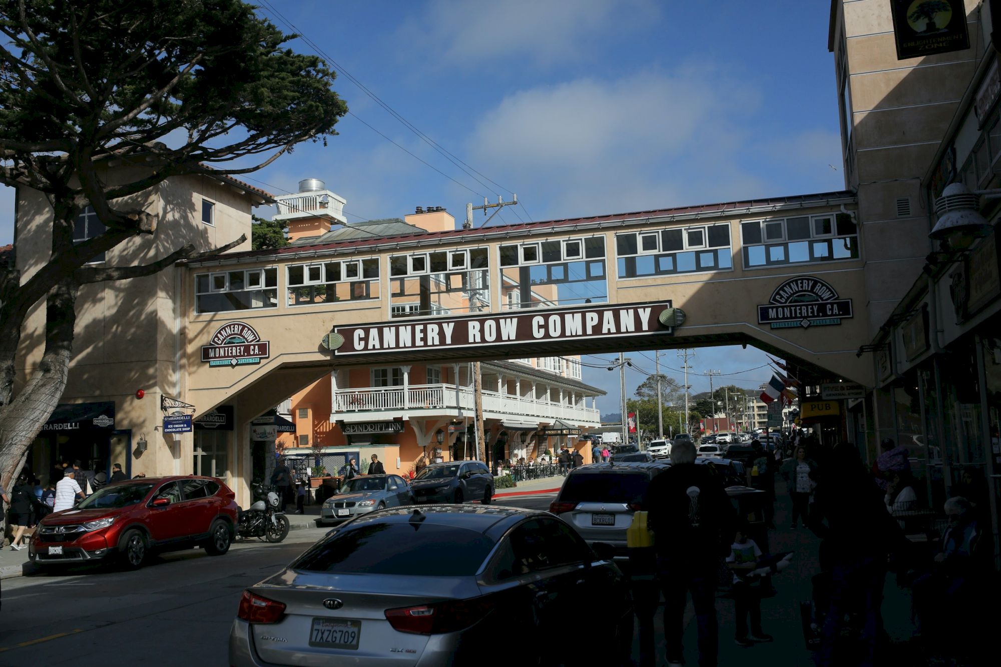 A busy street under a high archway that reads &ldquo;Cannery Row Company,&rdquo; with cars, people, shops, and blue sky in a coastal town.