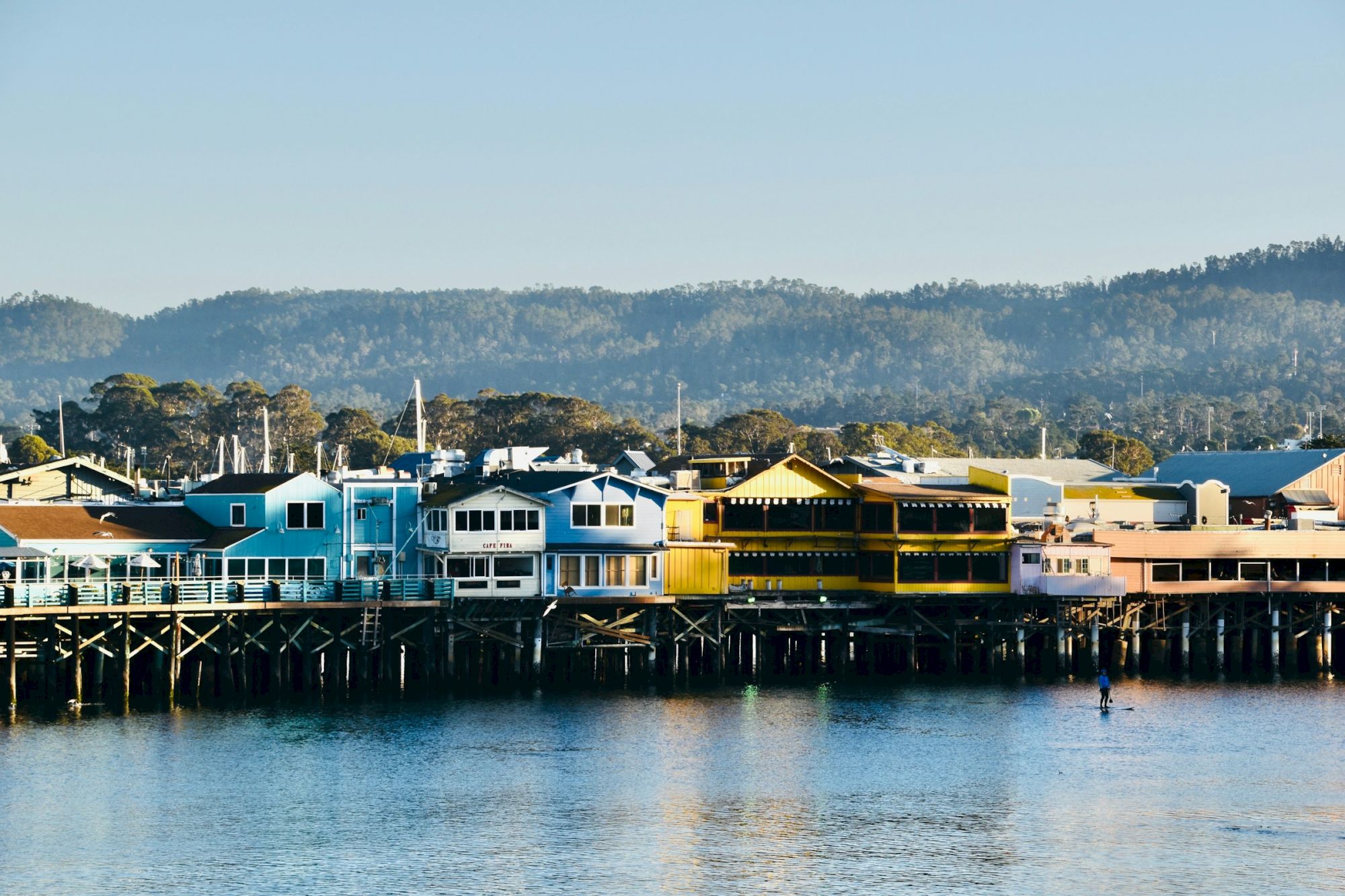 Colorful houses on a wooden pier by calm water, with hills in the background and clear blue sky, a peaceful coastal town scene.