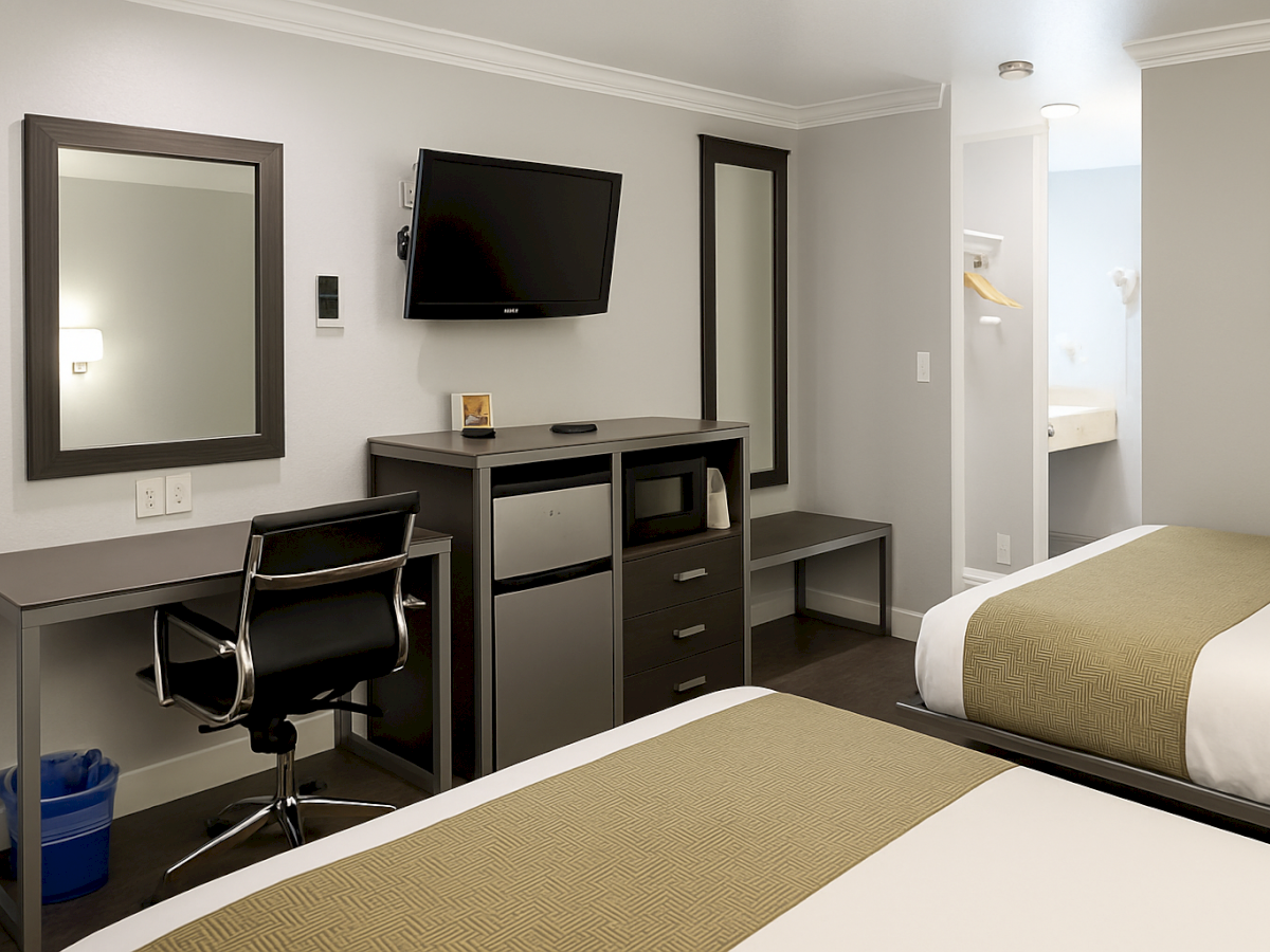 A modern hotel room with two single beds, a desk and chair, a wall-mounted TV, a mini-fridge and drawers, and a small bathroom doorway.